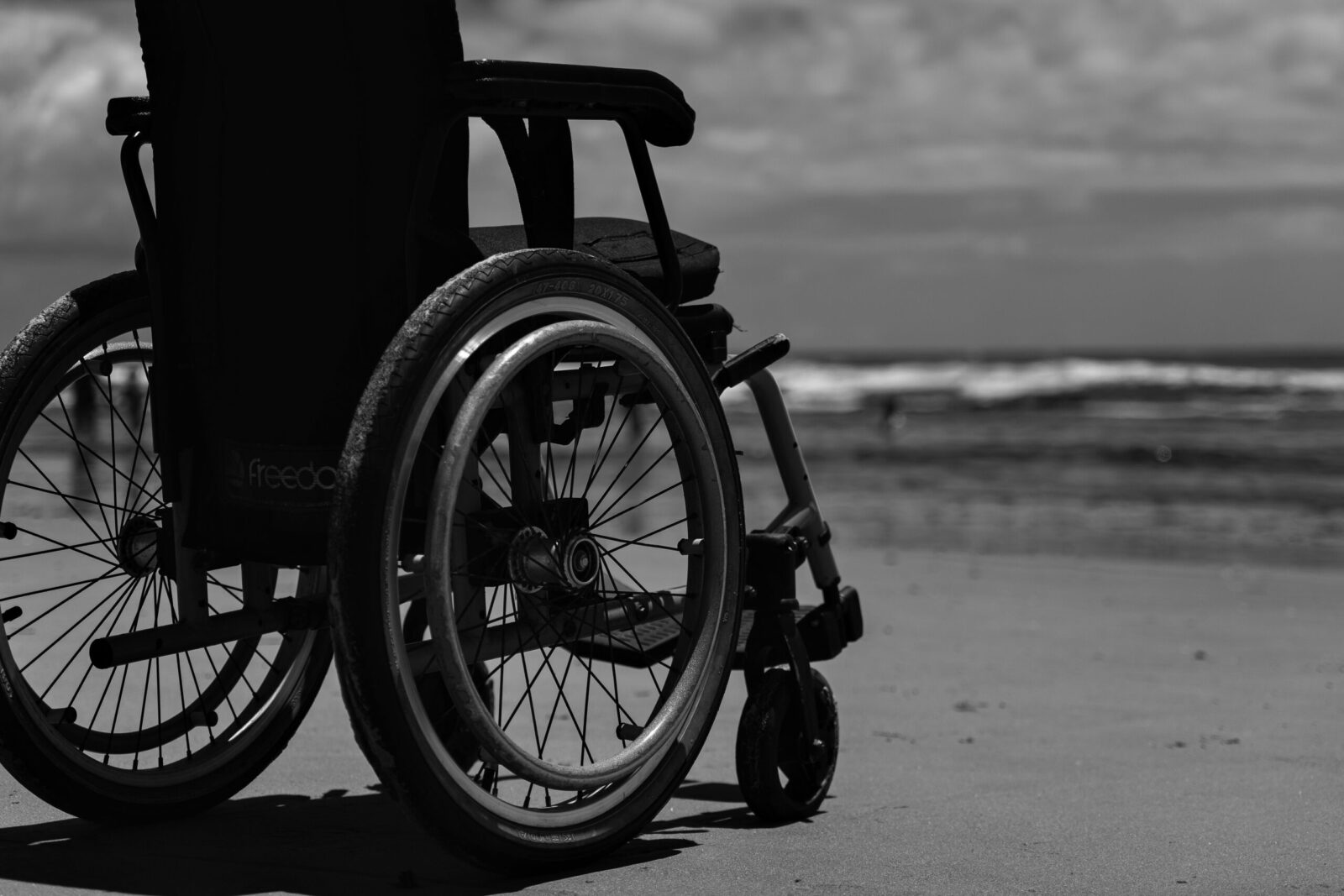 A monochrome image of a wheelchair on a sandy beach with ocean waves in the background.